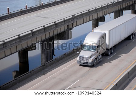 Big rig beige American bonnet long haul semi truck tractor transporting commercial cargo in dry van semi trailer running on overpass road along the river and upper level elevated highway