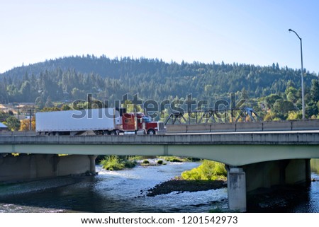 Classic red big rig semi truck with refrigerated semi trailer transporting commercial cargo and moving on the bridge across mountain river in stunning natural Columbia Gorge aria with wooded hills