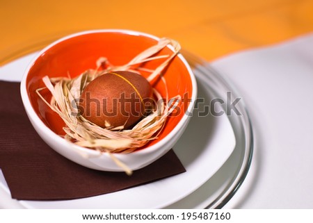 Brown striped Easter egg in orange bowl with straw on a background of napkins and other plates, white linen tablecloths and orange bedding