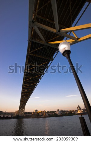 Modern perspective on the bridge over the river and city street lamp against sky and sunlit industrial area on beach with reflection in the water of the river and bridge structures illuminated edges.