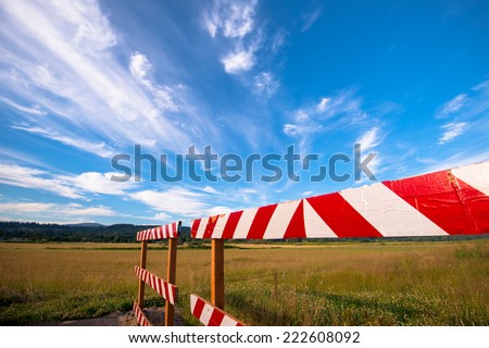 Fence with white and red striped ribbon, signifying the end of the road, which is on display at the border of the road in the middle of meadows, forest belt on the horizon and the clouds.