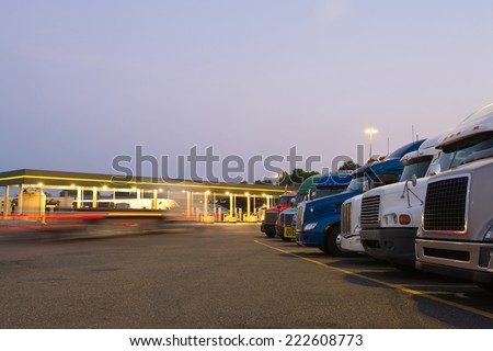 Number of semi trucks of various models and colors in the future on the night truck stop with a lit gas station and blurred lights of a passing semi truck and reflection of the lights on the big rigs.
