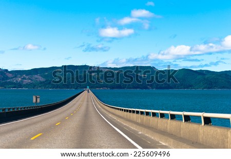 Long scenic road bridge with several lanes with raised sections and fencing in wide mouth of the Columbia River in Astoria, Pacific, rests on the horizon on the opposite bank of the hilly with trees.