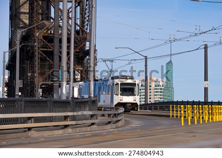 Modern white streetcar and power lines on turn from old drawbridge with rusty farm across river Willamette in Portland Down Town on background of modern office buildings of concrete and glass.