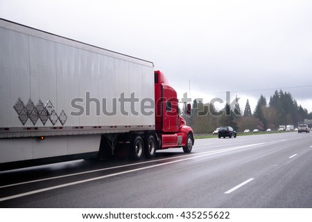 Beautiful stylish red big rig semi truck with a long covered dry van trailer with cargo moving for delivery to consumers on a wide multilane clear wet road after rain weather