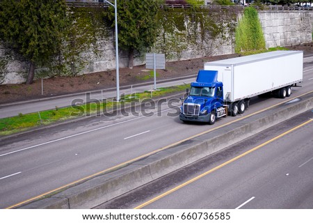 A blue semi truck with day cockpit and a roof spoiler to reduce air resistance and improve aerodynamics carries the trailer with cargo along the road in an urban city area