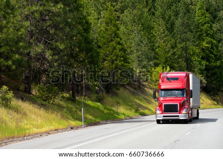 Modern popular for professional drivers model of semi truck in red going with dry van trailer transporting commercial cargo to destination warehouse by wide highway with green forest on background