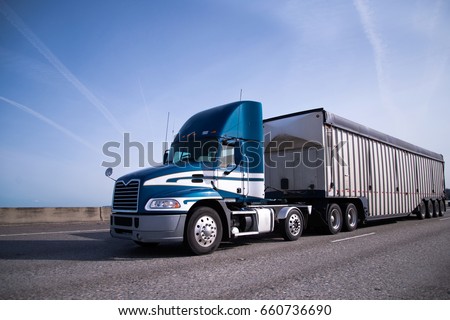 Contemporary big rig blue semi truck with high day cab and covered semi trailer for delivery bulk cargo moving on the wide highway with blue sky and little clouds on background