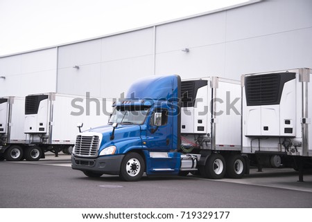 A big rig semi truck with a reefer trailer stands near the gate of the warehouse next to other reefer trailers that are loaded and unloaded to deliver perishable and frozen food to consumers