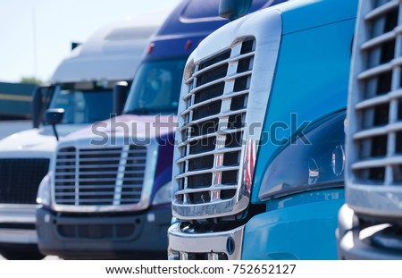 Grilles of different models of big rig semi trucks which stand in row on the truck stop parking waiting to continue delivery cargo