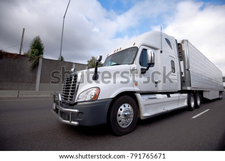 White modern American bonnet popular big rig semi truck for long haul delivery with reefer unit on refrigerator semi trailer running on the highway with concrete fence and cloudy sky