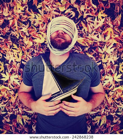 a man with a rope wrapped around his head laying in a pile of leaves holding a sailboat toned with a retro vintage instagram like filter effect