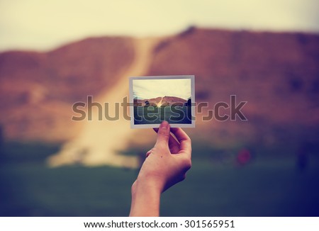 a young girl holding an instant photo like a polaroid in front of a landscape that is the same but a close up instead of a wide angle toned with a retro vintage instagram filter app or action effect