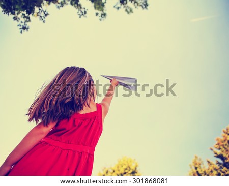 a cute girl in a red dress throwing a paper airplane into the sky while a real plane is passing overhead in a park during summer time toned with a retro vintage instagram filter app or action effect