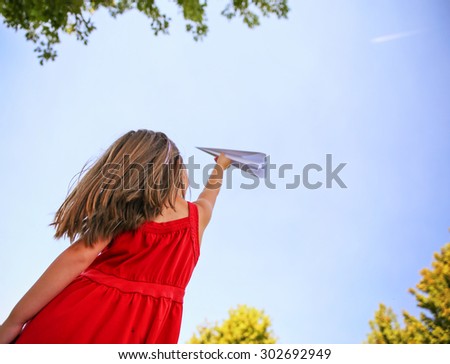 a cute toddler girl in a red dress throwing a paper airplane into the sky while a real plane is passing overhead in a park during summer time