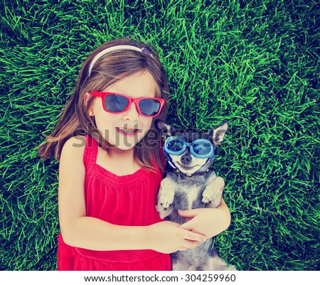 a cute toddler girl with red sunglasses on holding a chihuahua with goggles on in the grass in a park or backyard with a green lawn toned with a retro vintage instagram filter effect app or action