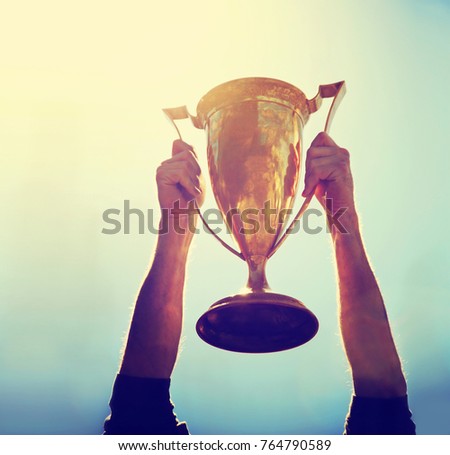 a man holding up a gold trophy cup as a winner in a competition toned with a retro vintage instagram filter effect app or action (SHALLOW DOF on writing)