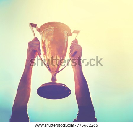 a man holding up a gold trophy cup as a winner in a competition toned with a retro vintage instagram filter effect app or action (backlit with the sun)