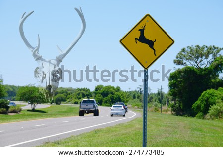 Deer Crossing sign by road with cars and a deer skull ghosted into the background representing the dangers for wildlife crossing roads.