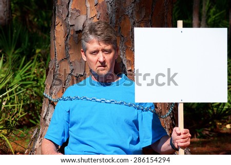 Tree hugger eco warrior man holding up blank sign and he is chained to a tree to protest deforestation while trying to save this tree\'s life