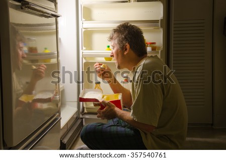 A man who has nighttime sleep-related eating disorder sleep eating as he sits in front of a refrigerator eating ice cream out of carton, oblivious to everything around him as he is actually sleeping.