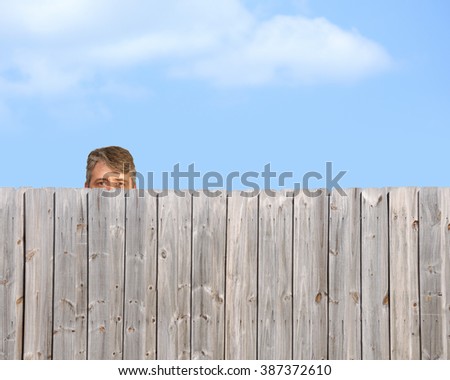 A goofy, happy man is being a peeping tom and nosy neighbor by stalking, watching and gawking over a wooden privacy fence