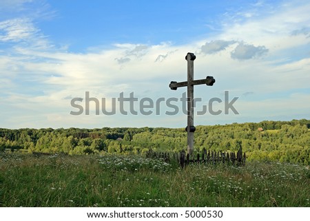 Christian wood cross on grass overgrown hill (and forest colony far away)