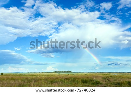 Rainbow in blue cloudy sky above summer meadow.