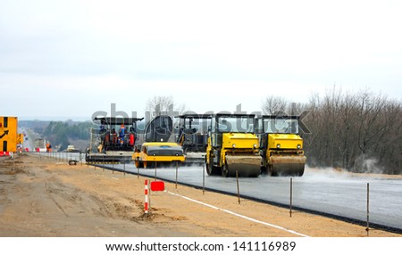 Road crew operate automatic asphalt spreader and steam roller follows