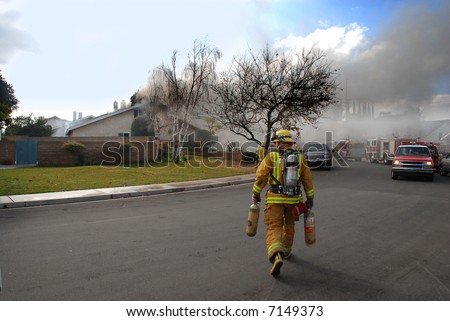 Fire fighter carrying air tanks heads to a house on fire with fire fighters on the roof.