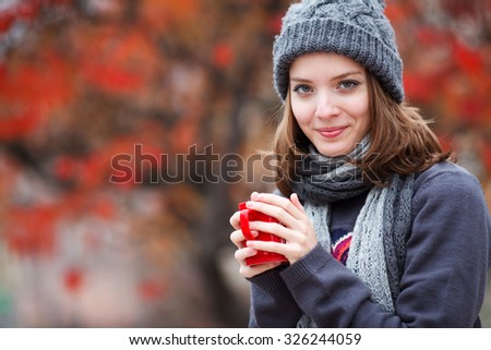 Fall concept - autumn woman drinking coffee on park bench under fall foliage. Beautiful young modern woman smiling happy and cheerful