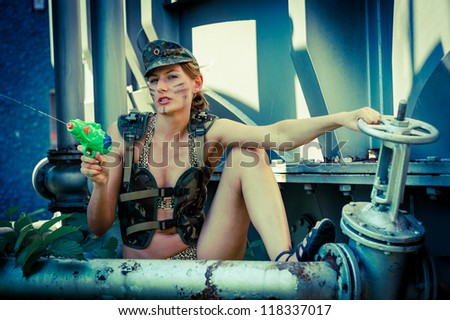Woman in military camouflage shoots from a water pistol on the industrial background