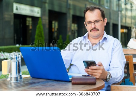Middle age businessman works on laptop and messaging on a smartphone in outdoor cafe