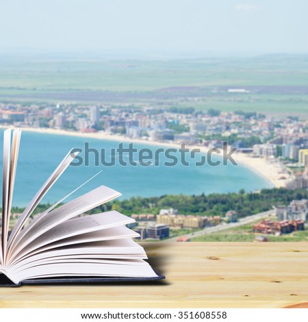 Open book on a rustic wooden table. In the background, a resort town and the sea