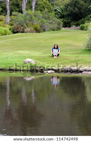 Woman reading newspaper sitting in grass with a pond in the foreground