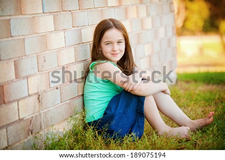 A young girl outdoors leaning up against a brick wall.   Plenty of negative space on the brick wall.  She is happy and relaxed in a jeans skirt and blue top