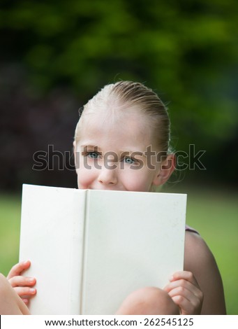 A happy young 7 year old girl with blond hair and blue eyes is looking over a white book outdoors.  She is studying or reading a novel.