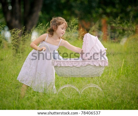 A pretty little girl plays with her doll in a vintage pram in the long grass.  She is wearing a classic white dress.  The vibrant green color of the long grass frames the girl.