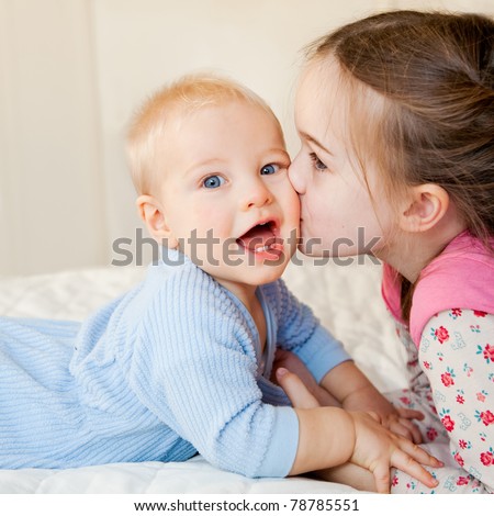 This is a sweet moment between a big sister and her baby brother.  The baby is looking at the camera with a look of delight.  The sister is kissing his cheek.