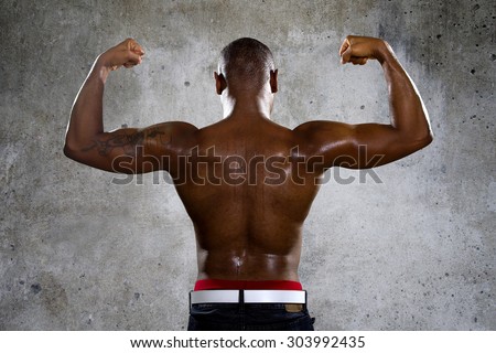 Fit black man in hip hop style clothing flexing back muscles. He is shirtless to show off his muscles and has a tattoo on one arm.