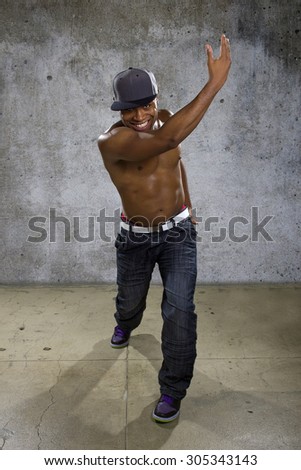 Shirtless muscular black man wearing urban hip hop style clothing on concrete background.  He is sweaty and physically fit.  The man is topless to show off his muscles.