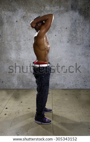 Shirtless muscular black man wearing urban hip hop style clothing on concrete background.  He is sweaty and physically fit.  The man is topless to show off his muscles.