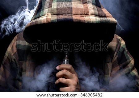 Man with concealed identity smoking a controversial vape.  Vaping is debatable in the health community if it is safe or a health risk.