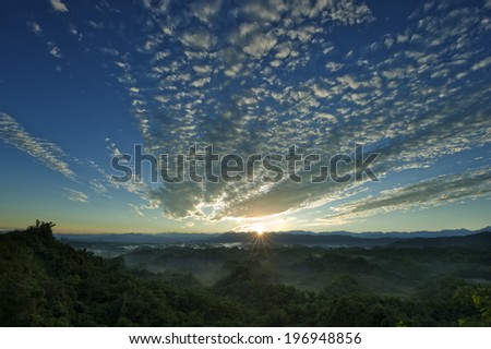A cloudy sky and sun above tree covered mountains.