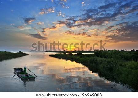 A person on a boat in a narrow body of water and sunrise in the distance.