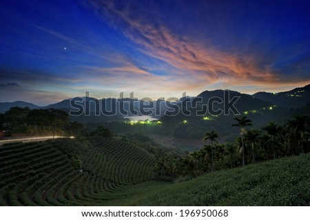 A tropical hilly area with green lights in the distance under a cloudy sky.