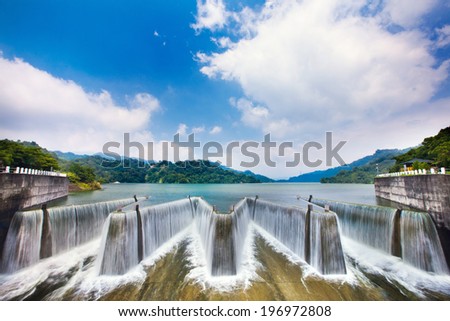 Blue sky partially filled with fluffy white clouds, water falling over dam structures in the foreground.