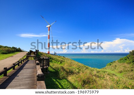 A narrow road beside a wood walkway with a wind turbine and water in the distance.
