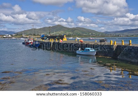 Old stone harbor wall at Knightstown, Valentia Island, Kerry, Ireland, with colorful leisure and fishing boats moored by the pier. Fluffy white clouds and blue sky are reflected in the calm sea water.