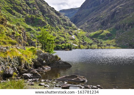 Sunlight sparkles on a peaceful lake near the Gap of Dunloe, Killarney, Ireland, and a steep narrow path, the tourist route taken by jaunting cars and hikers, leads up through the mountain pass.
